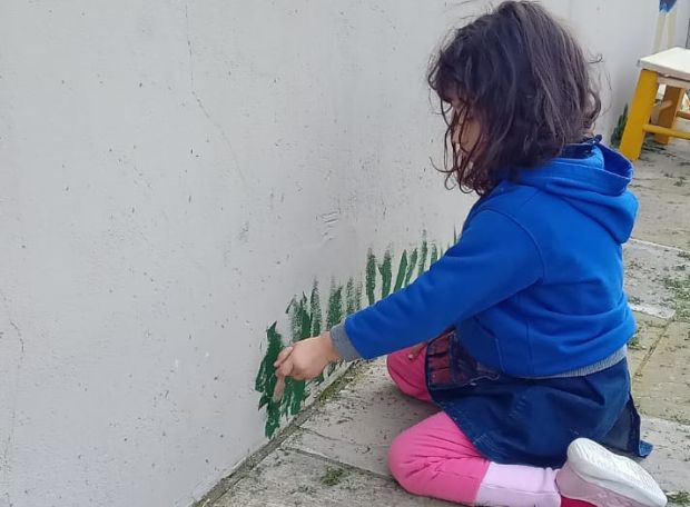 Niña pintando con pincel sobre una pared blanca durante una actividad artística al aire libre.