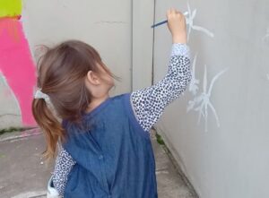 Niña pintando con pincel sobre una pared durante una actividad artística al aire libre.